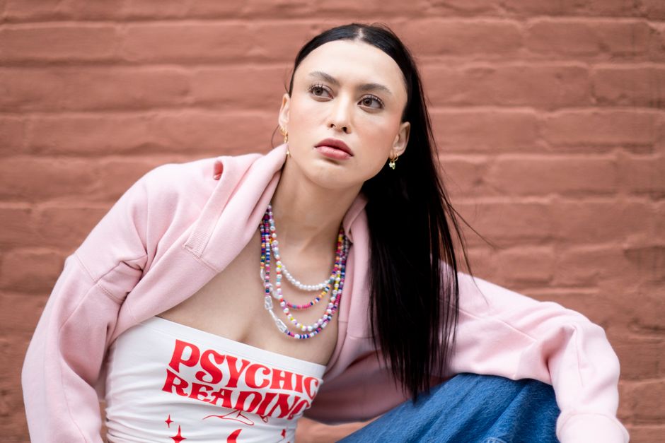 Stylish woman in a pink jacket with layered necklaces posing against a brick wall.