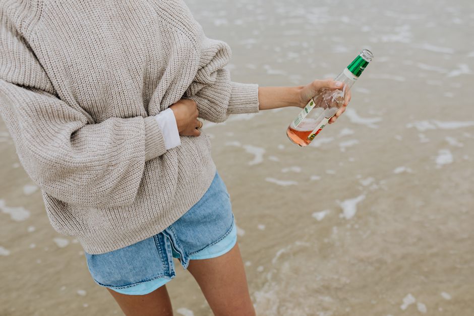 A woman holds a beverage bottle while strolling on the beach in casual summer attire.