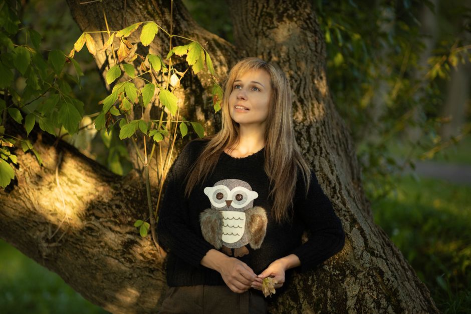 Woman in a cozy owl sweater leaning against a tree in a forest setting, enjoying nature.