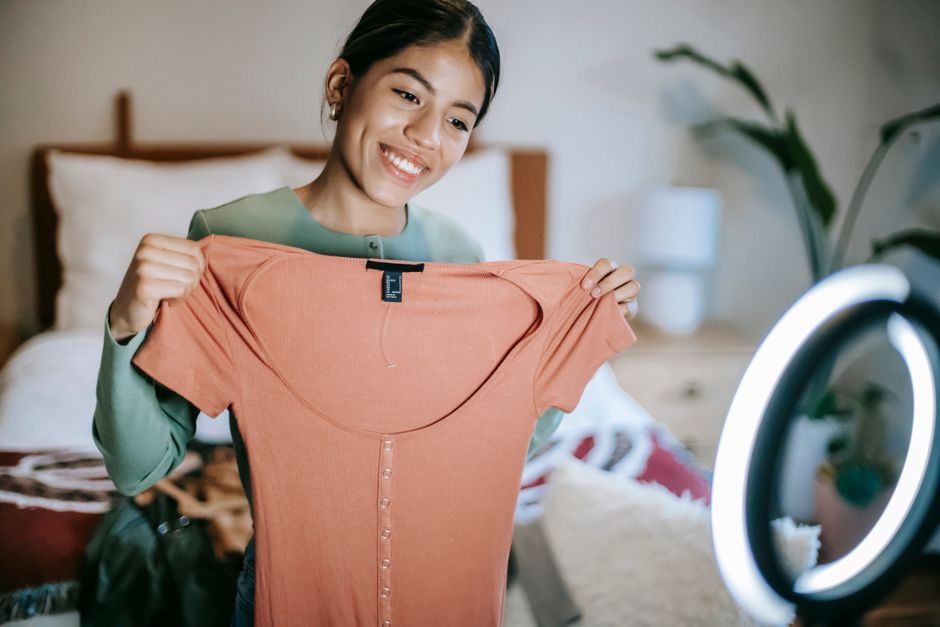 Happy young ethnic female in casual outfit showing pink dress while selecting clothes near ring lamp and bed near potted plant while standing in light room at home