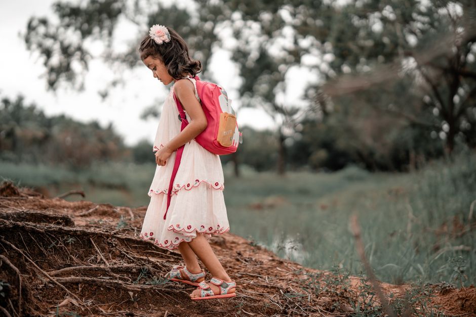 A young girl in a white dress and pink backpack hiking through a forest trail.