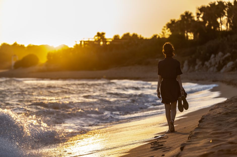 A woman strolls along a sandy beach during a golden sunset, holding her shoes.