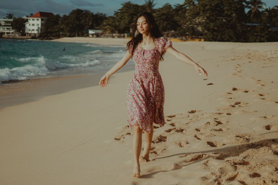 A barefooted woman in a floral dress enjoying a sunny day on the beach, with footprints in the sand.