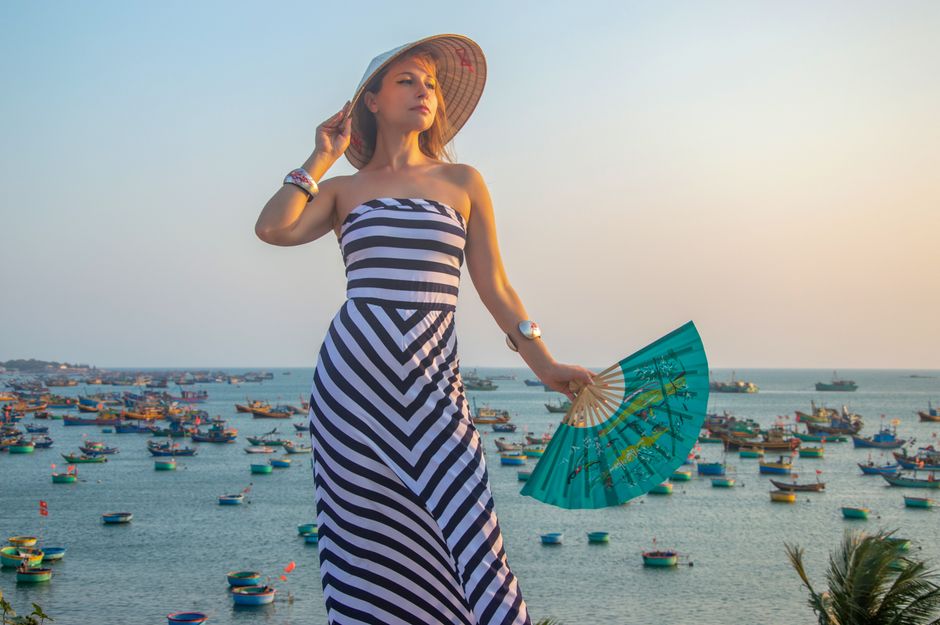 Stylish woman in striped dress with fan stands by a colorful sea full of boats at sunset.