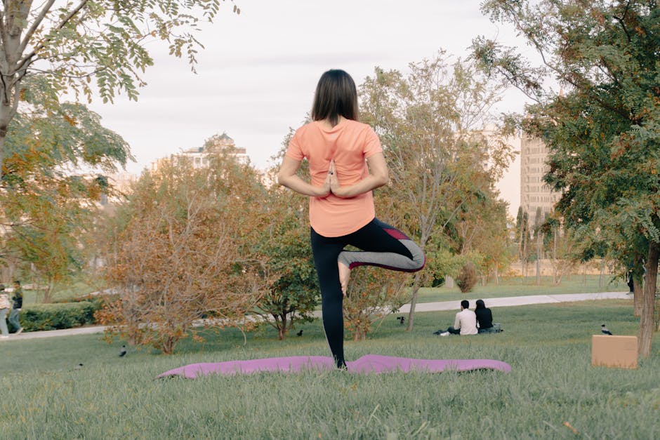 Adult woman performing yoga on a mat in a park, surrounded by greenery and trees.