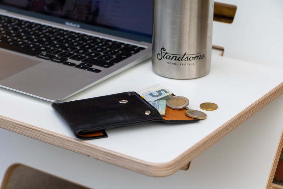 A sleek workspace featuring a laptop, wallet with euros, and a stylish metal tumbler on a minimalist desk.
