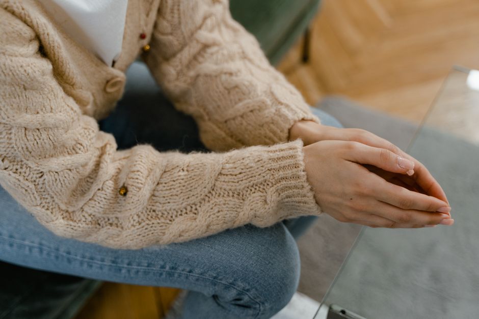 A person in a knitted sweater with folded hands sitting indoors on a couch.