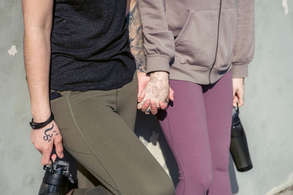 Crop anonymous lesbians with tattoo and bottles of water in activewear holding hands while standing near wall on sunny street