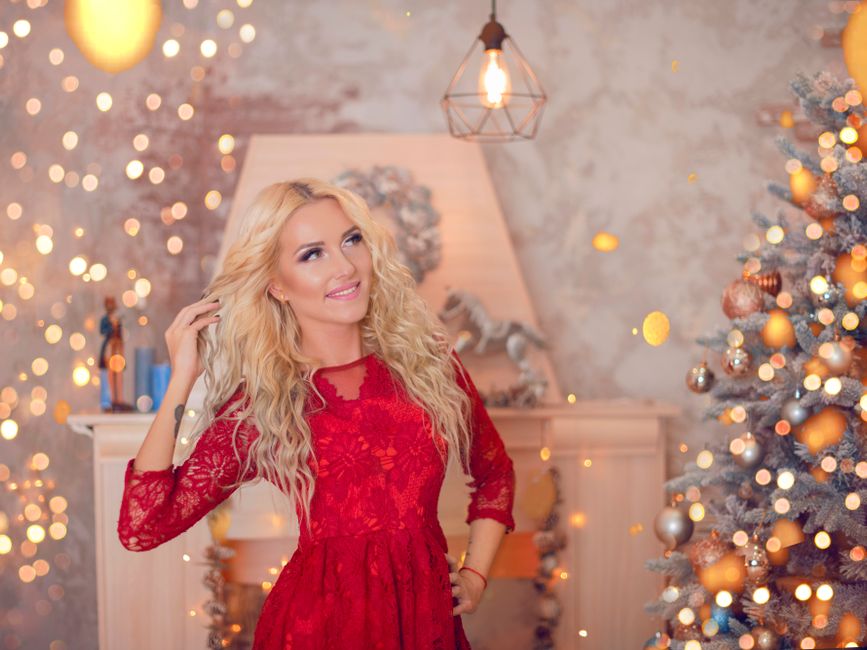 Woman in a red dress joyfully posing beside a decorated Christmas tree indoors.