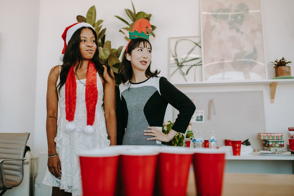 Two women enjoying a festive office holiday party with Santa hats and red cups.