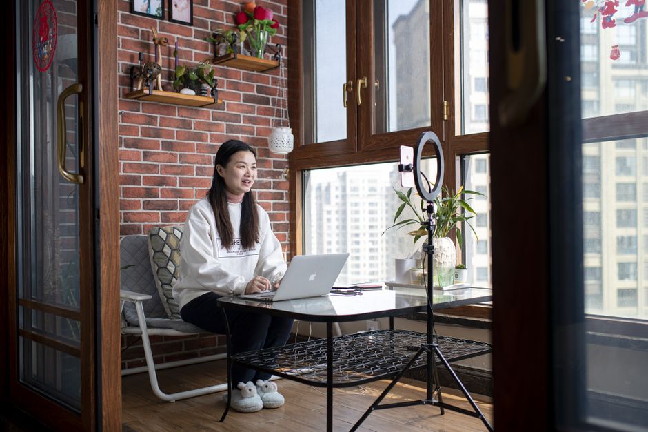 Woman working remotely in a sunlit Wuhan apartment with laptop and ring light setup.