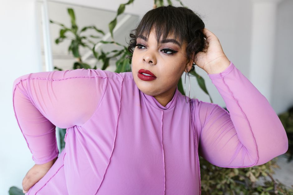 Confident plus-size woman with hand on head in a purple outfit indoors.