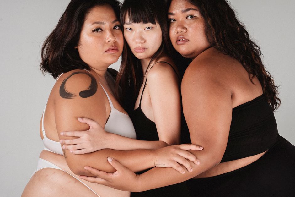 Calm Asian female models with different body constitution hugging each other while looking at camera on white background in studio