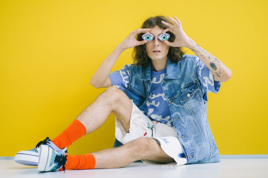 Young adult in fashionable denim and vibrant socks posing with eye props against yellow backdrop.