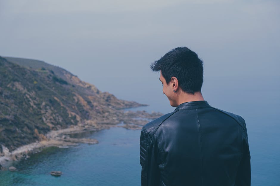 Rear view of a man in a black leather jacket gazing at a scenic coastal cliff and serene ocean.