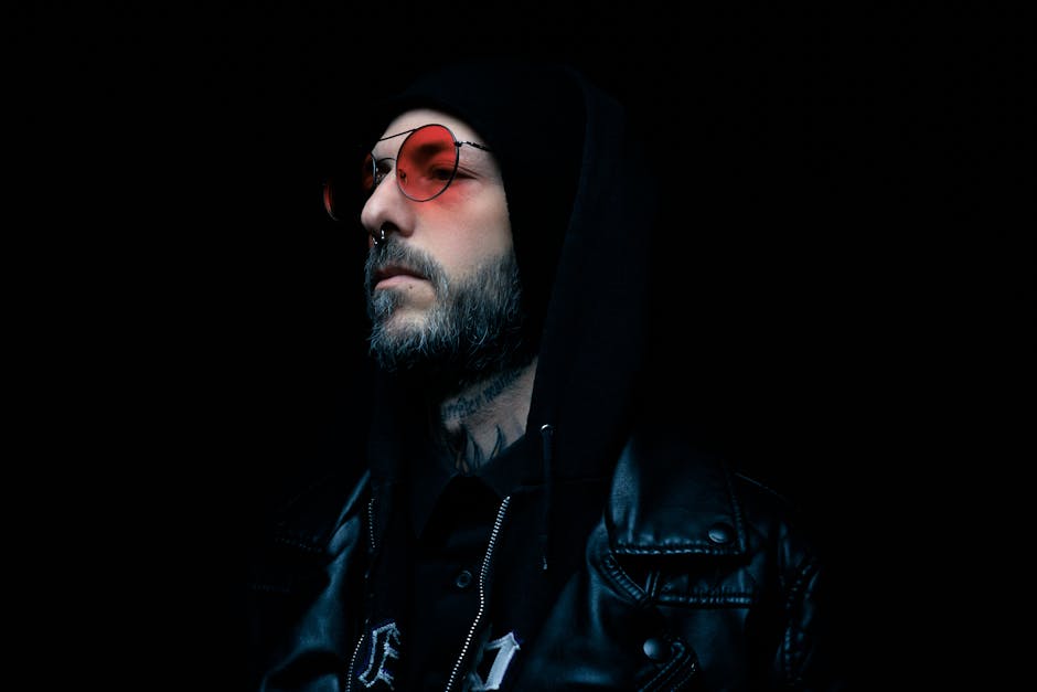 Moody portrait of a man in a black leather jacket and red tinted glasses with a dark background.