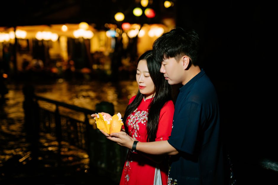 Couple holding a lotus lantern by the riverside, glowing under night lights.