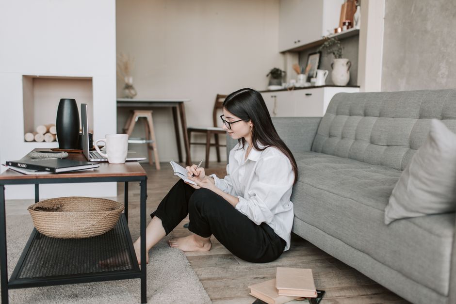 Side view of thoughtful young woman in eyeglasses and casual outfit sitting on floor near sofa and writing in notebook while resting in comfortable apartment