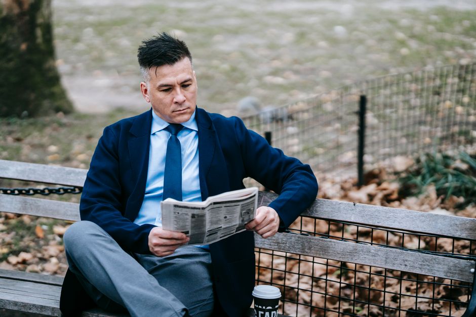Smartly dressed man reading a newspaper on a park bench, sipping coffee.