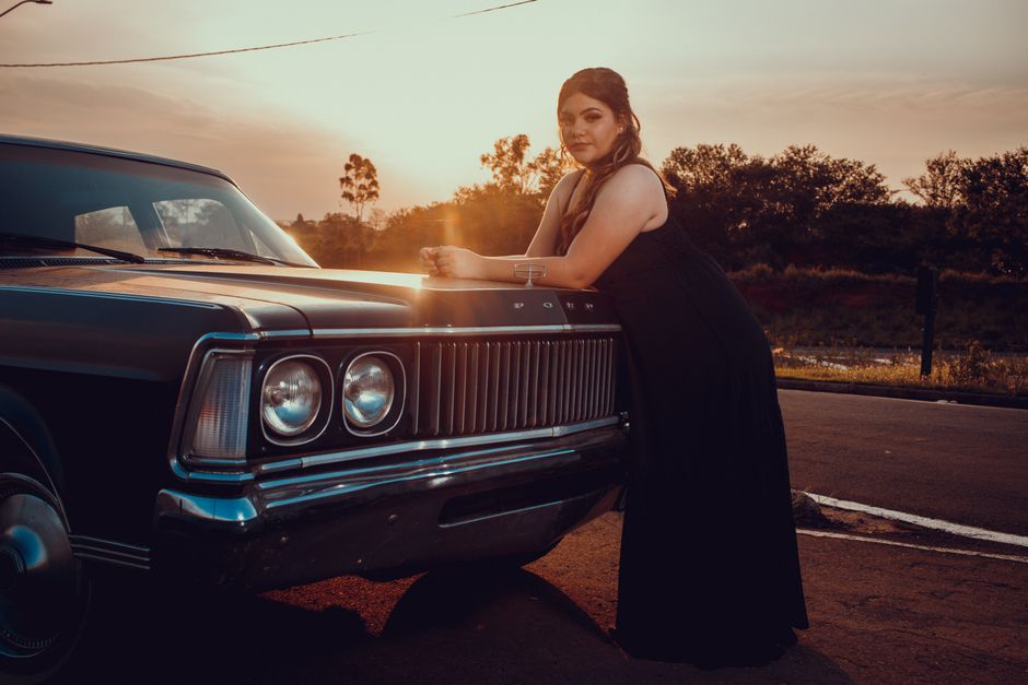 A woman in a black dress leans on a vintage Ford car during sunset on a scenic road.