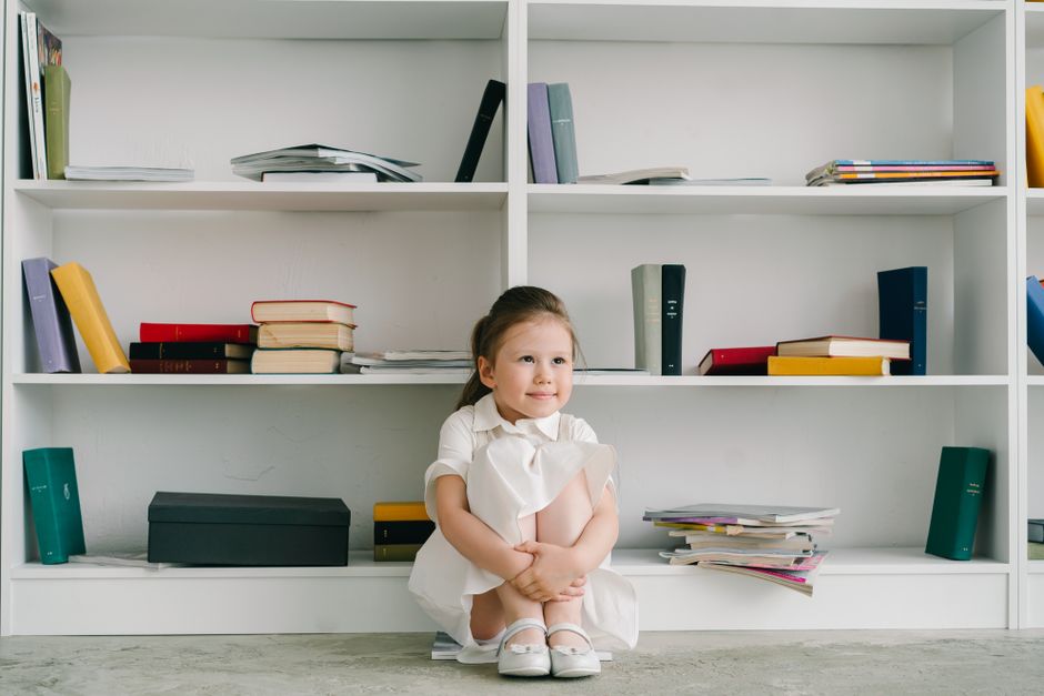 A cute child sitting on the floor by a bookshelf, surrounded by books in a cozy indoor setting.