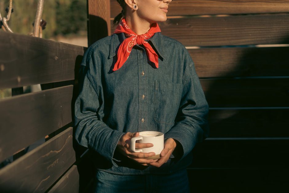 Woman wearing denim and a red bandana holding a coffee cup outdoors in the sunlight.