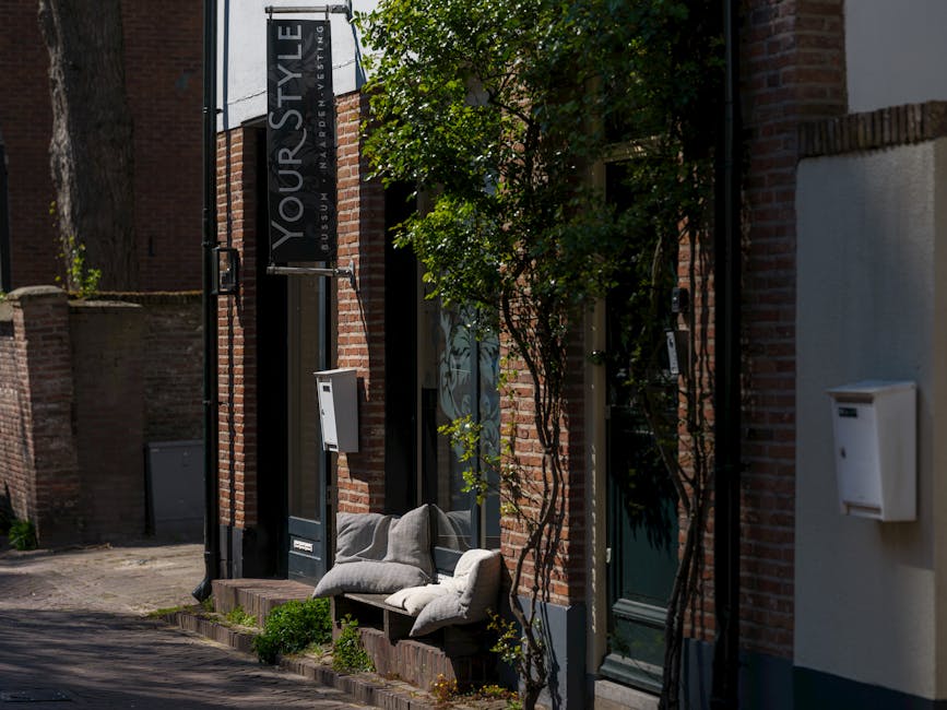 Outdoor seating with pillows outside a stylish brick-walled salon in an urban setting, enveloped by greenery.