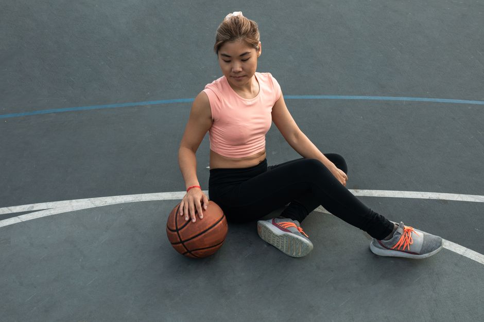 East Asian woman in sportswear sitting on an outdoor basketball court, holding a basketball.