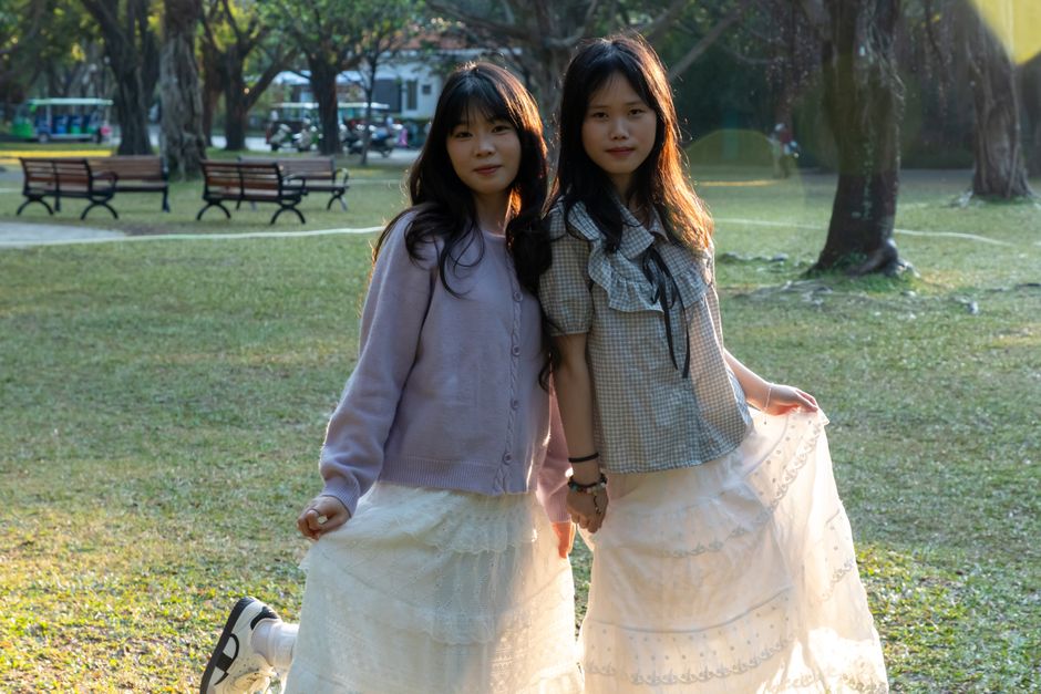 Two young women enjoy a sunny day in the park, posing in stylish outfits with gentle backlighting.