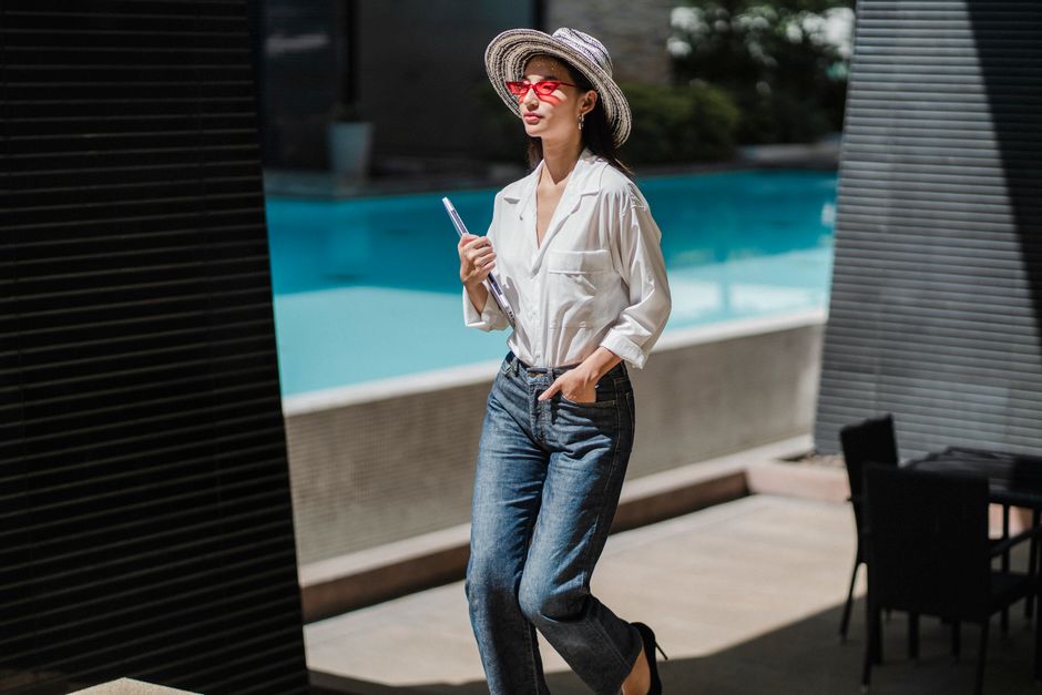 Self assured young ethnic lady in elegant outfit and sunglasses walking near outdoor swimming pool with laptop in hand during summer vacation