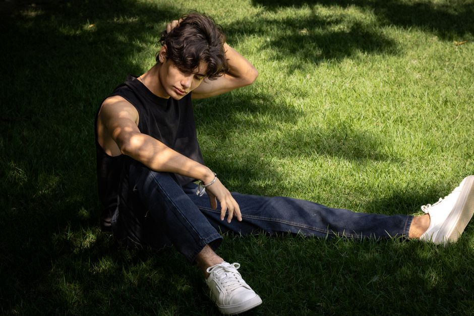 A young man in casual attire sits thoughtfully on a sunlit grass lawn.