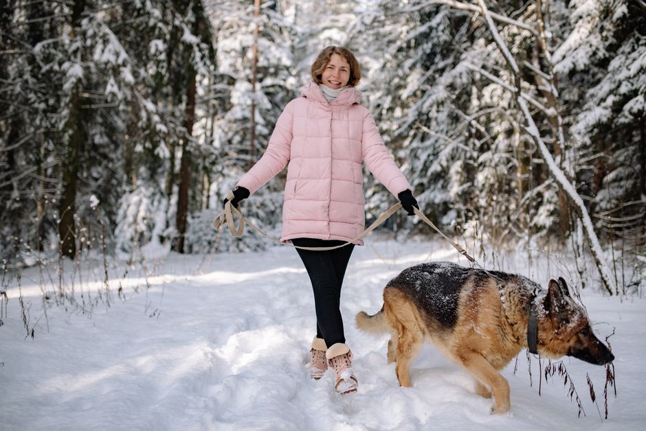 A woman in a pink coat walks a German Shepherd dog in a snowy forest during winter.