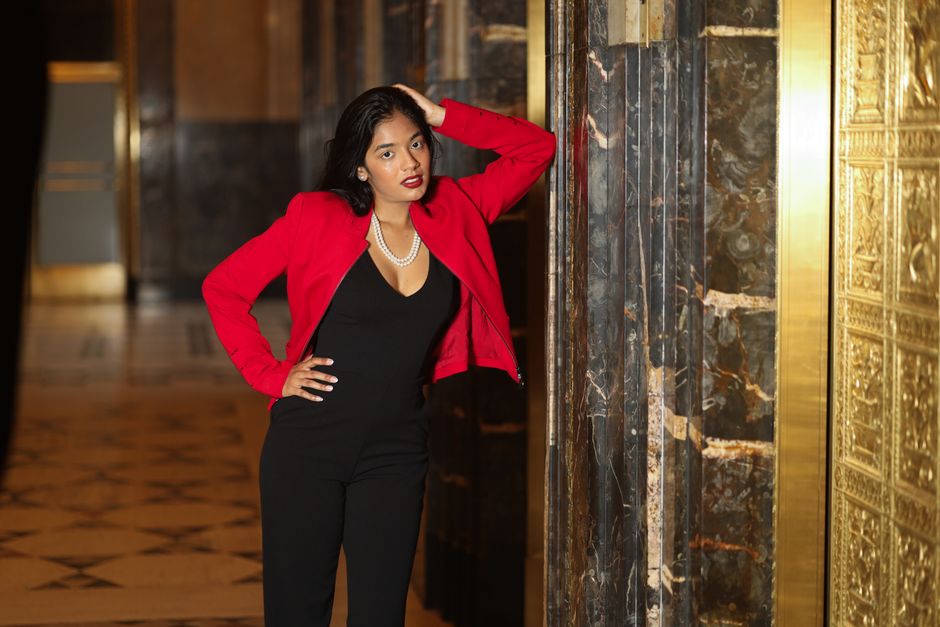 Stylish woman in red jacket posing elegantly in a luxurious indoor setting.